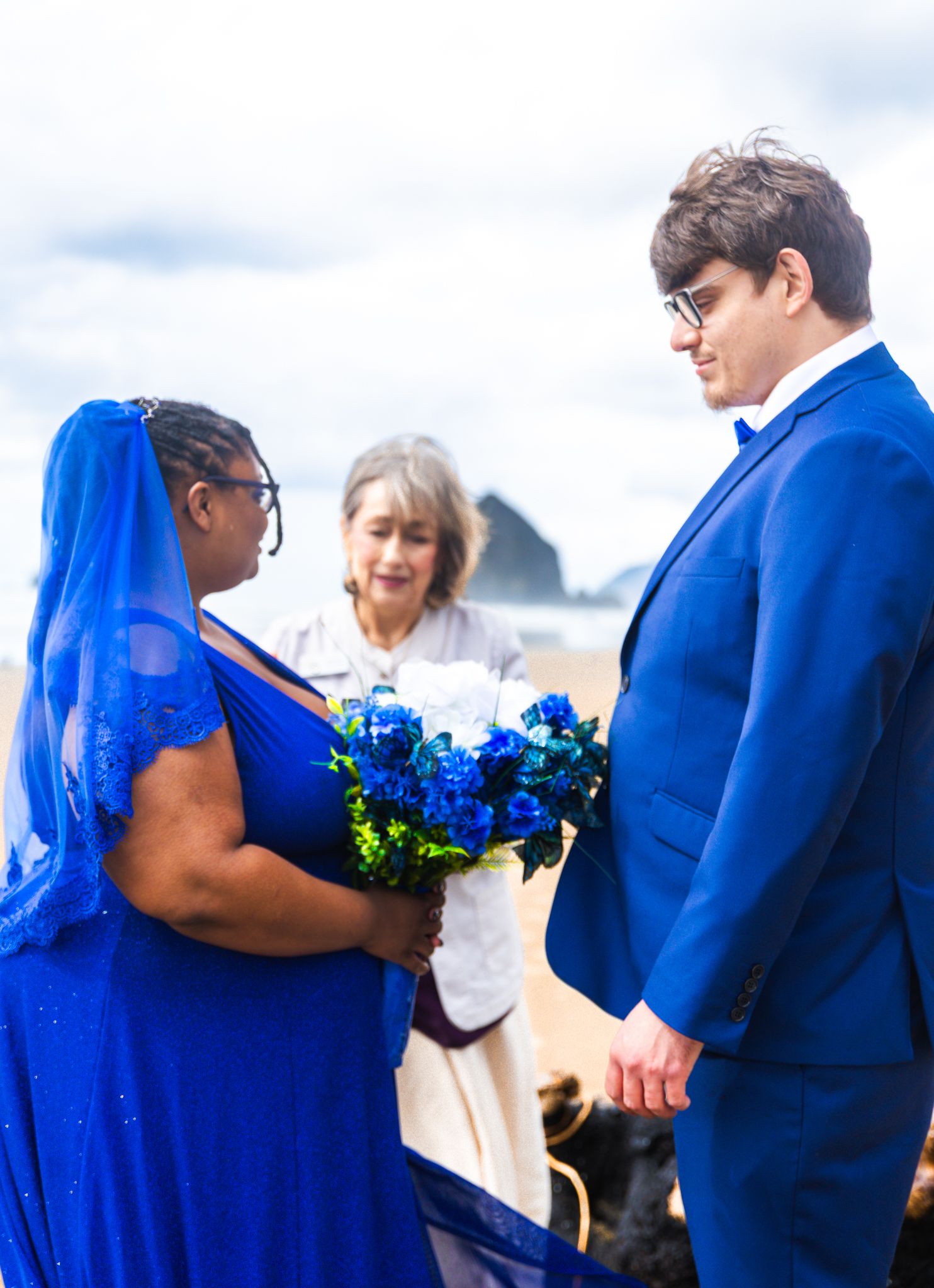 Couple in matching blue outfits exchange vows on beach with officiant, bride holding blue floral bouquet.