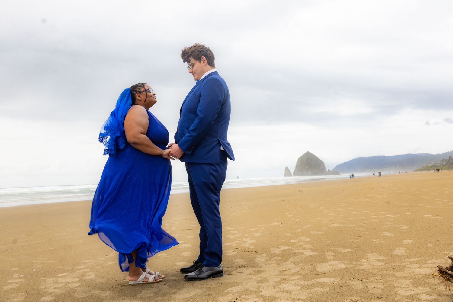 Couple in matching blue outfits holding hands on a sandy beach with Haystack Rock in the background.
