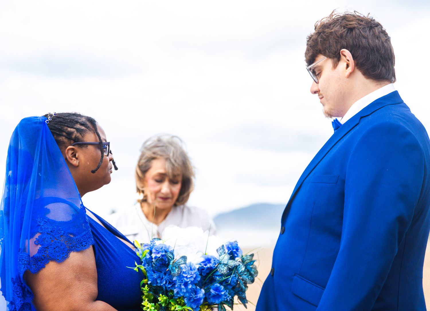 Bride and groom in matching royal blue attire exchange vows with officiant outdoors at a wedding ceremony.