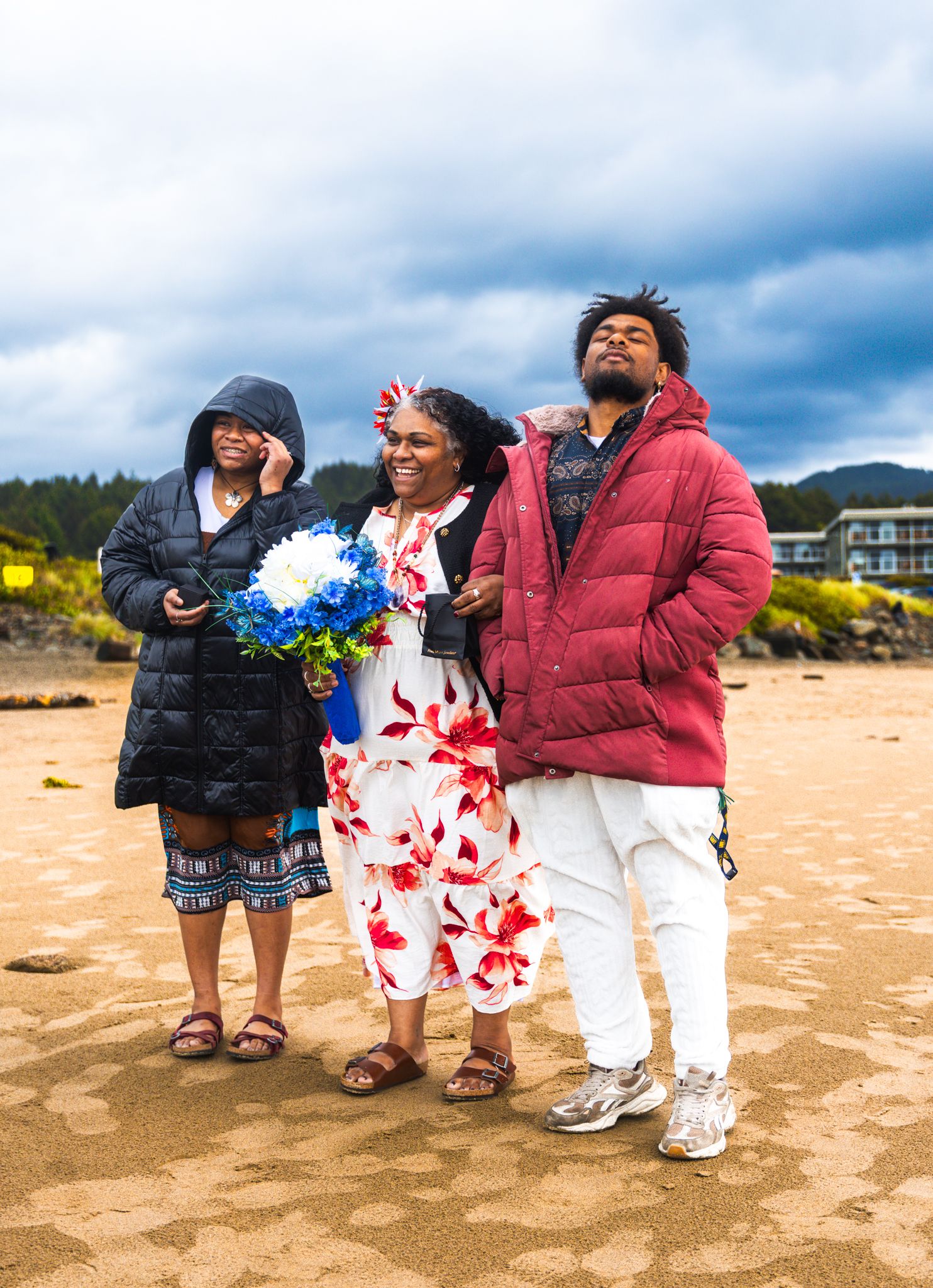 Three people pose on a sandy beach on a cloudy day, with one woman holding blue and white flowers.