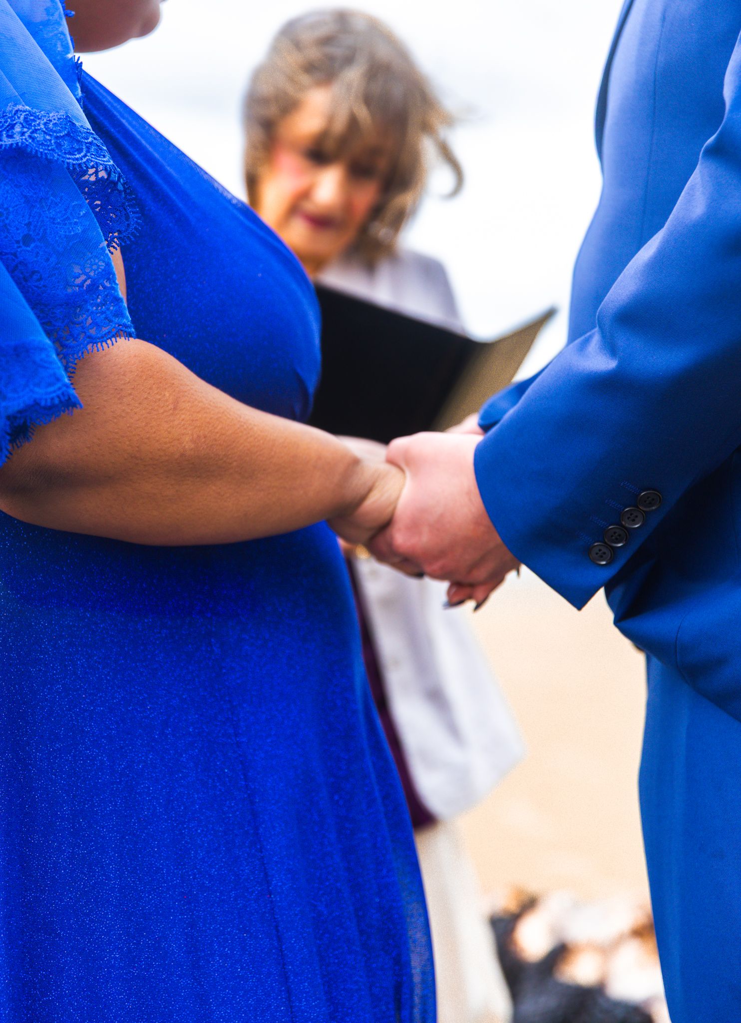 Two people in blue outfits hold hands during a wedding ceremony while an officiant reads from a book in the background.