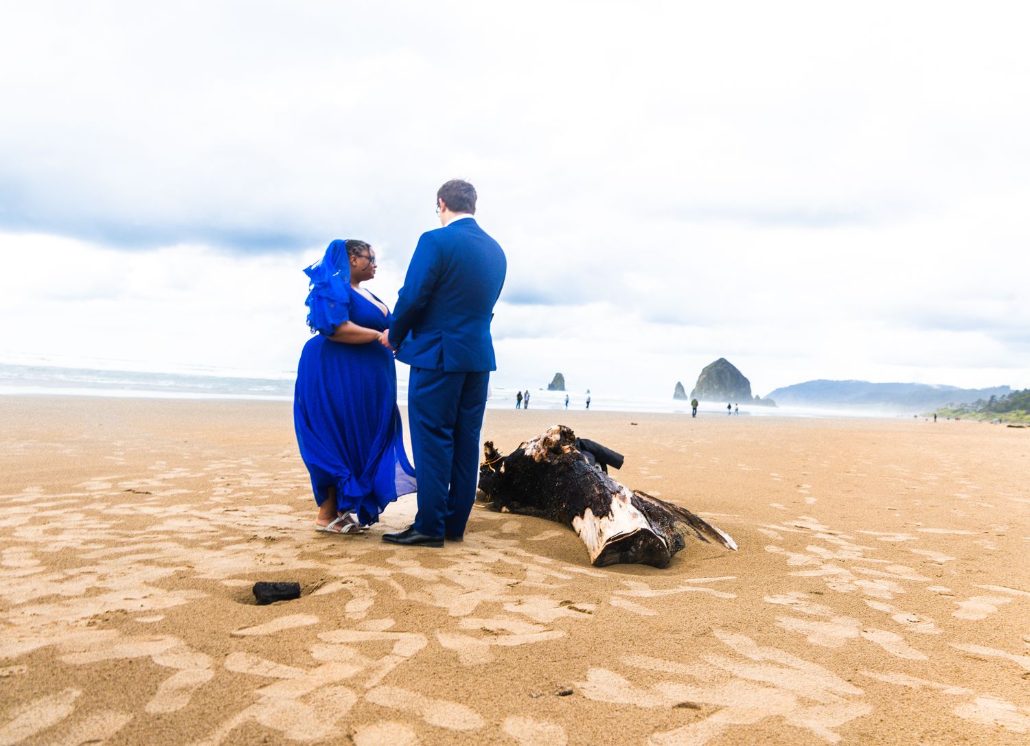 Couple in blue attire sharing a romantic moment on a sandy beach with Haystack Rock in the background.