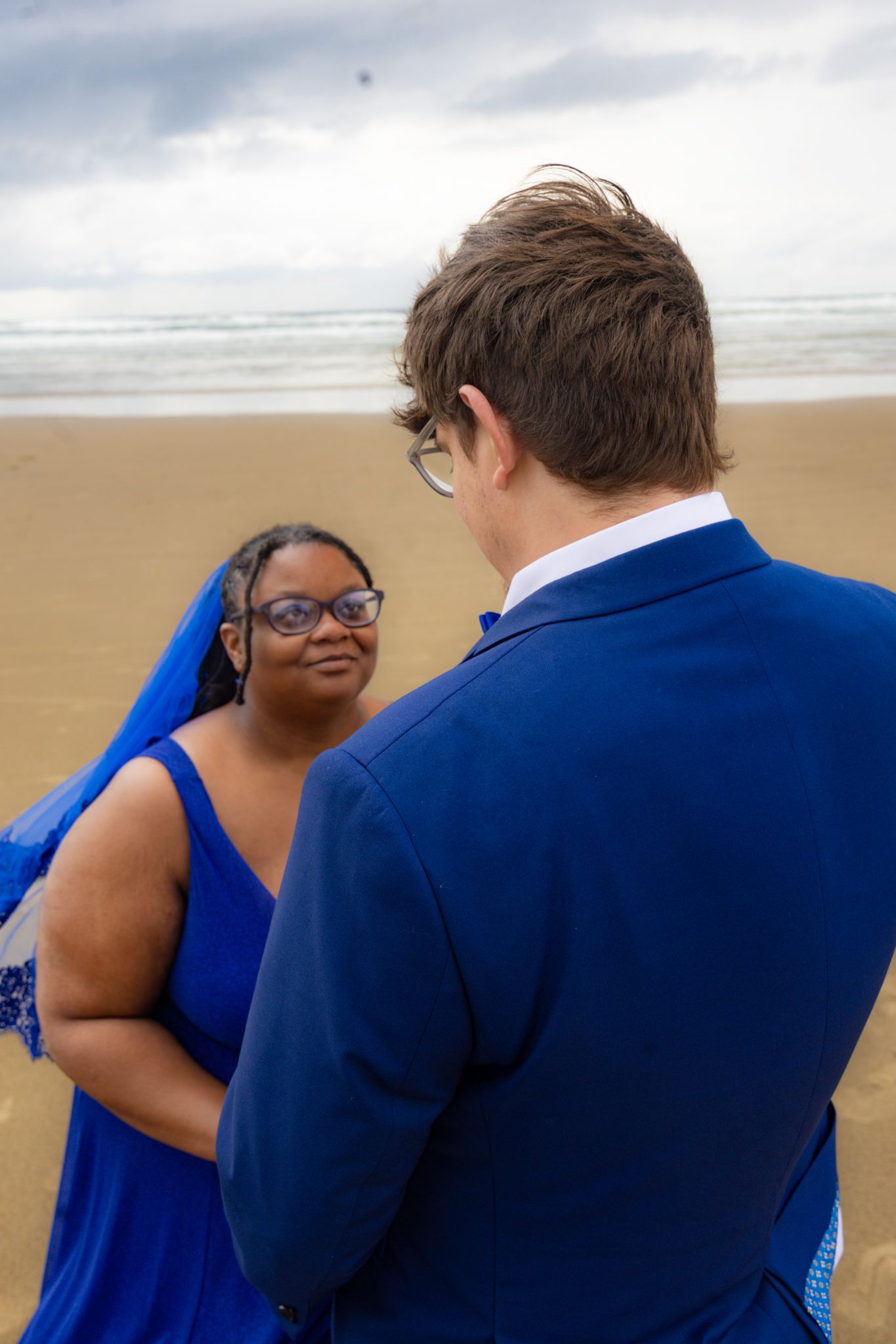 Interracial couple in matching blue outfits exchanging vows on a sandy beach with ocean backdrop.