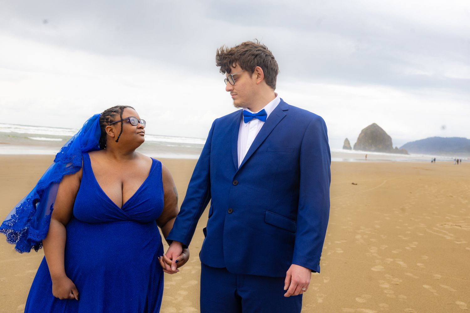 Couple in matching royal blue wedding attire holding hands on a sandy beach with Haystack Rock in the background.