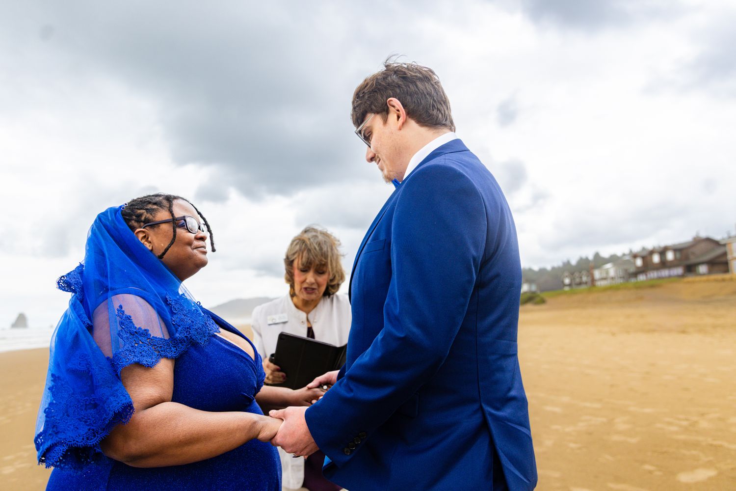 Beach wedding ceremony with couple in blue attire exchanging rings, officiant presiding under cloudy sky.
