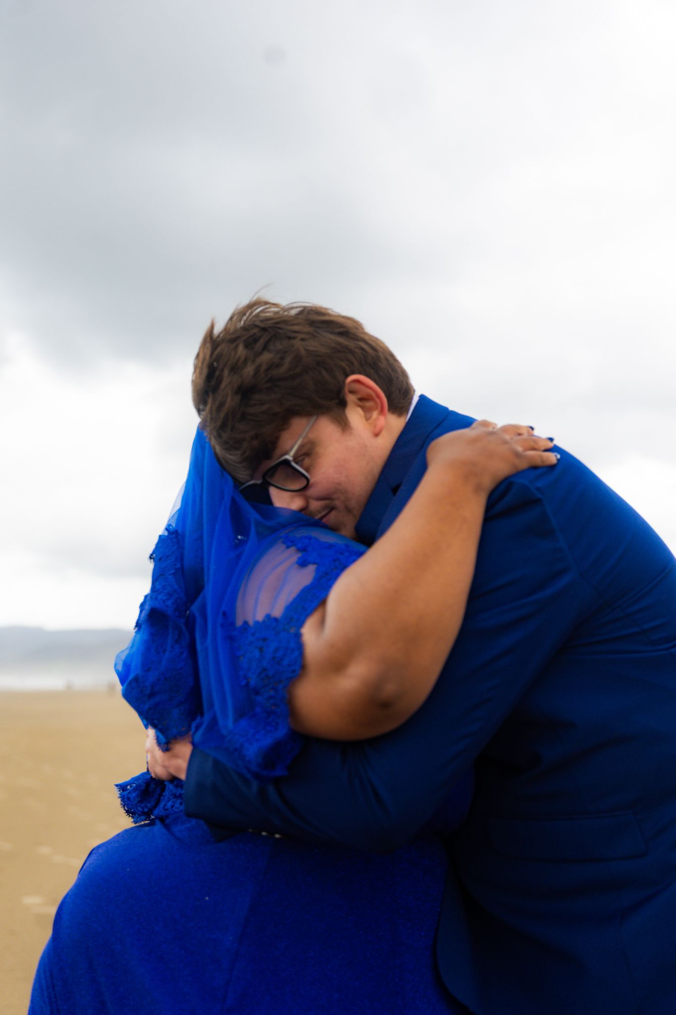 Two women in matching royal blue dresses share an emotional embrace on a cloudy beach.