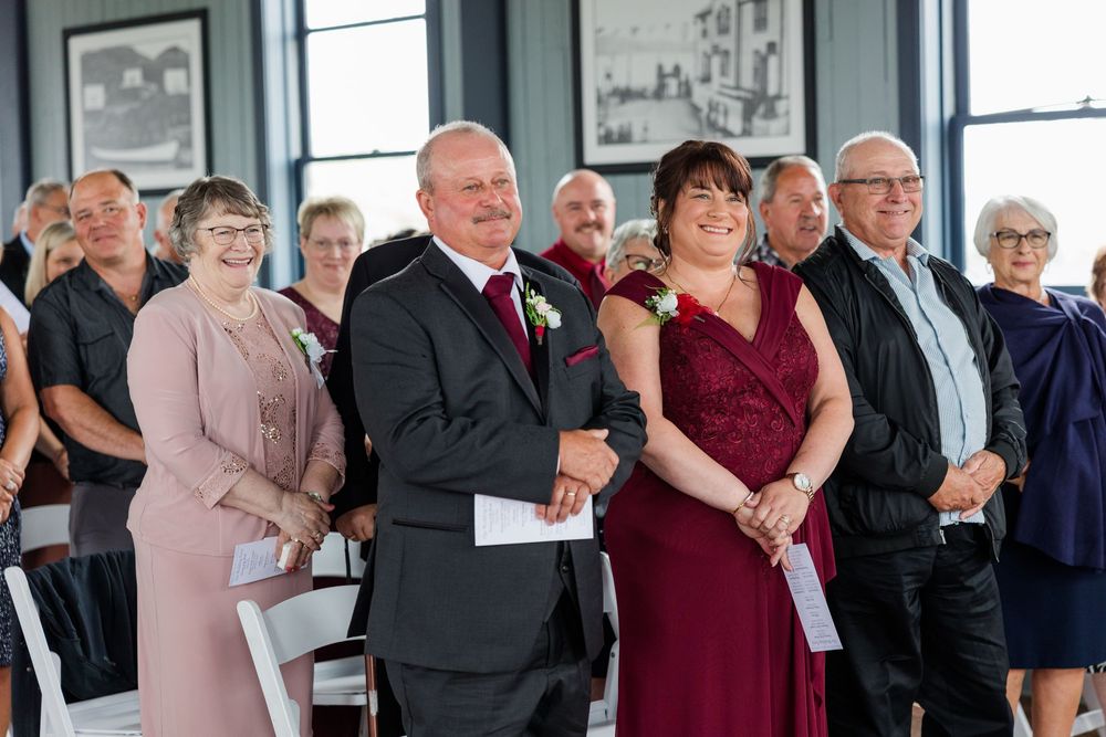 Guests smile during a wedding ceremony in a bright venue, with the wedding party in burgundy and black attire.