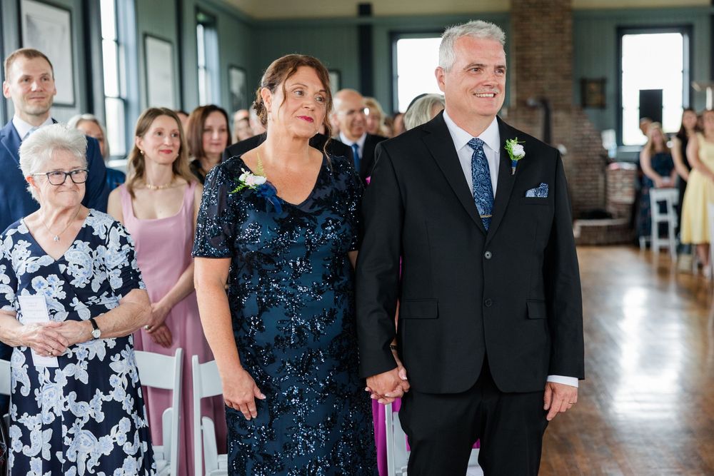 Couple in formal attire walking down wedding aisle, woman in blue lace gown, man in black suit, guests seated behind them.