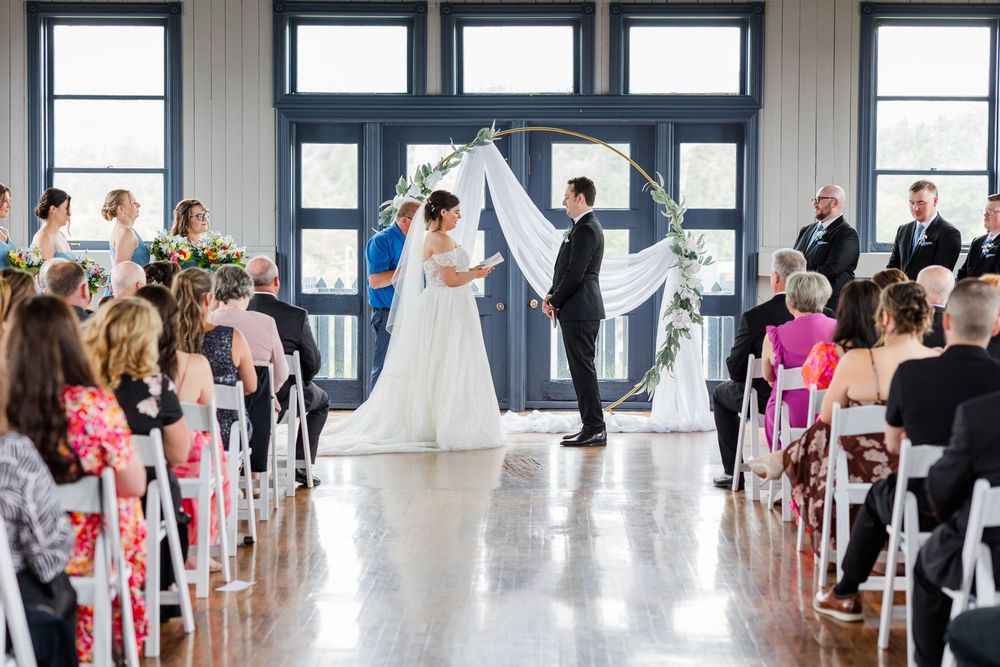 Elegant indoor wedding ceremony with bride and groom at floral arch altar, guests seated in white chairs.