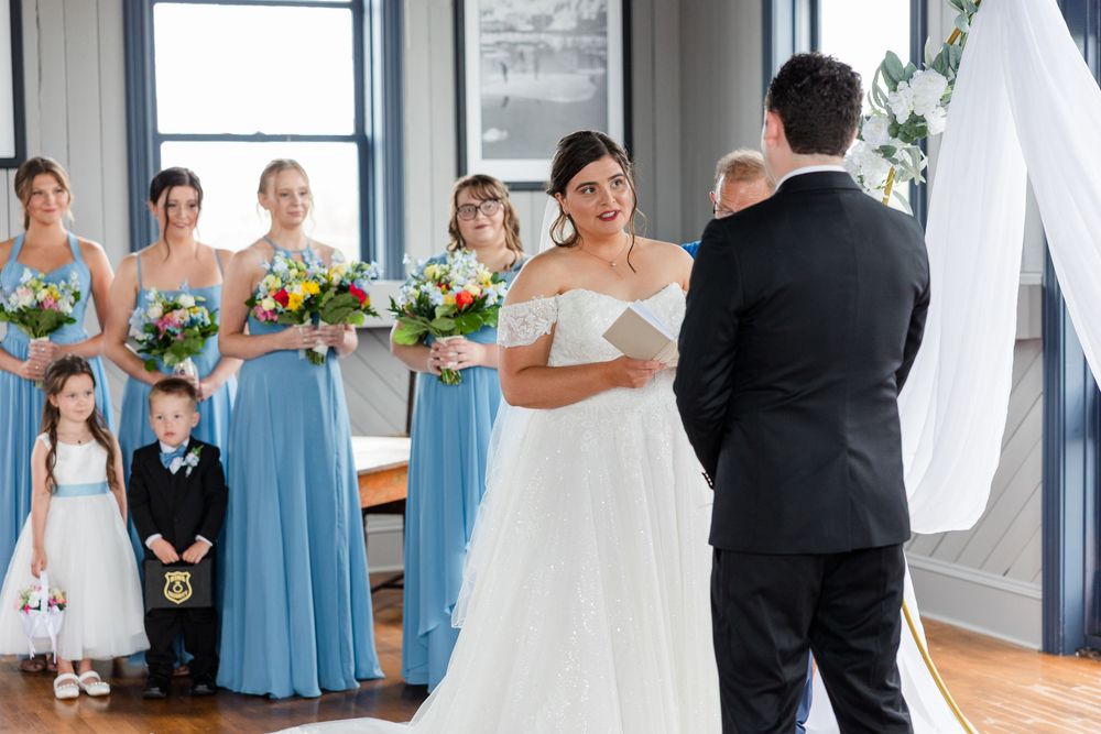 Bride in white gown reads vows to groom at indoor wedding ceremony with bridesmaids in blue dresses.