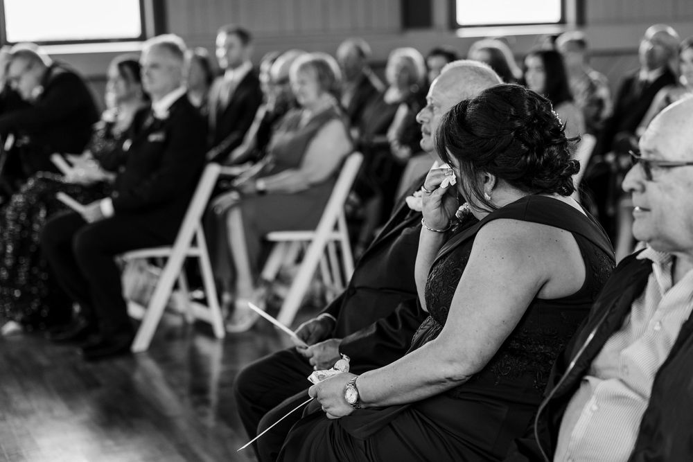Black and white wedding ceremony photo of emotional guests wiping tears while seated in white chairs.