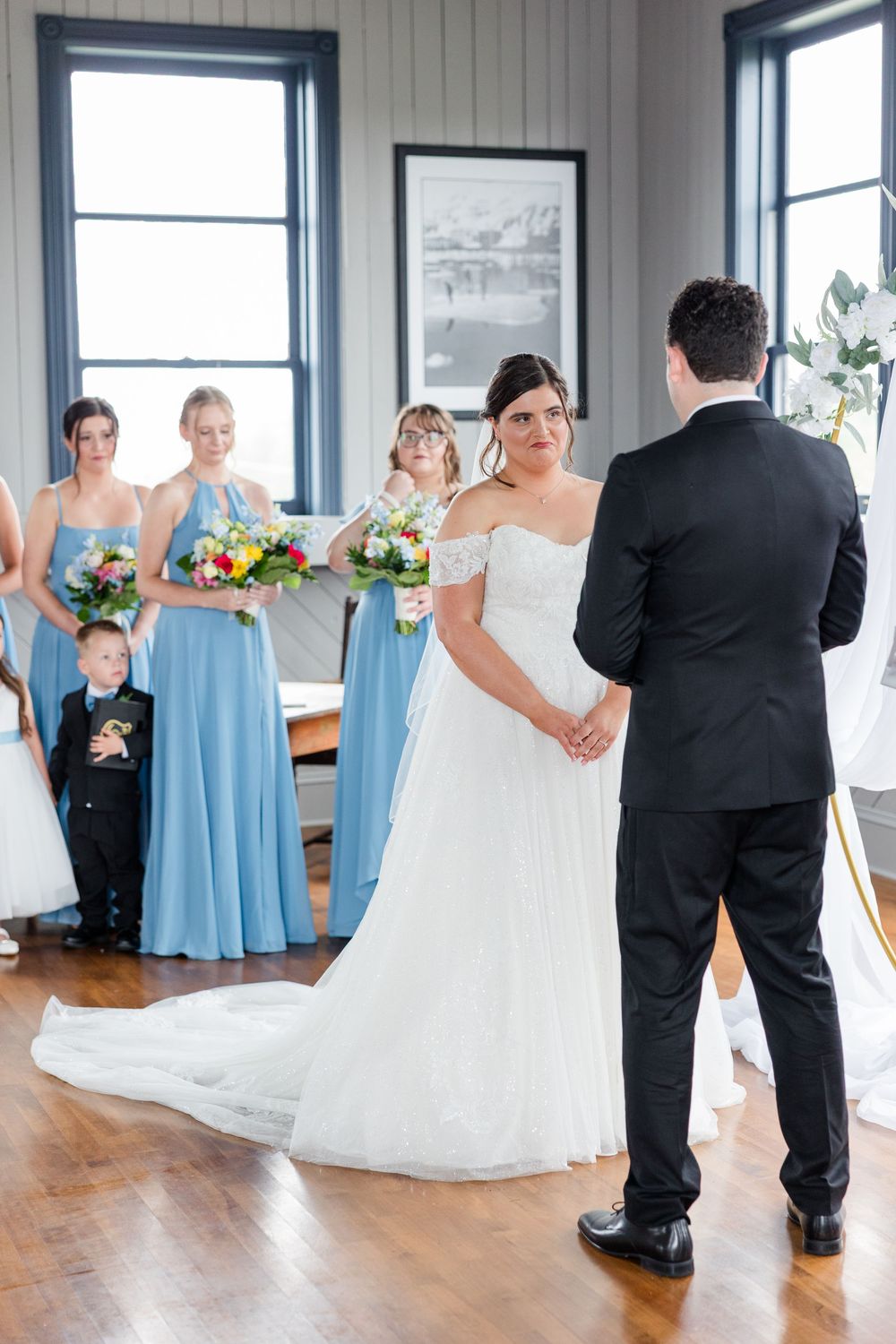 Bride and groom exchange vows indoors, with bridesmaids in blue gowns holding colorful bouquets standing behind them.