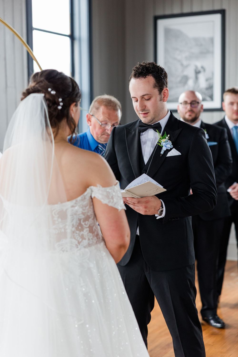 Groom reads wedding vows from a booklet to his bride during an indoor ceremony, with officiant and groomsmen behind.
