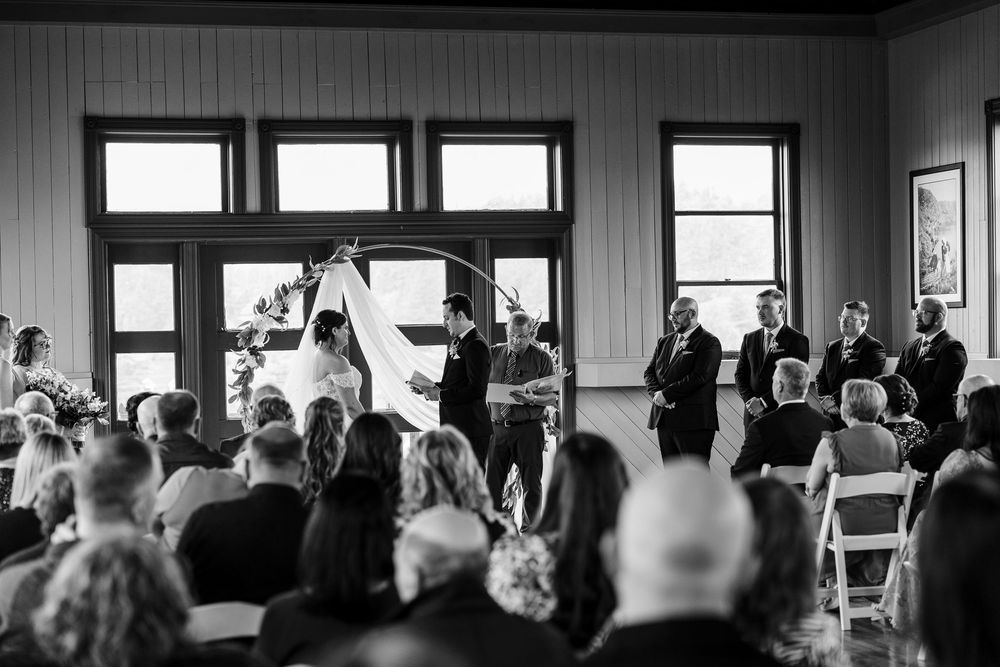 Black and white wedding ceremony photo with bride and groom at altar surrounded by guests in elegant indoor venue.