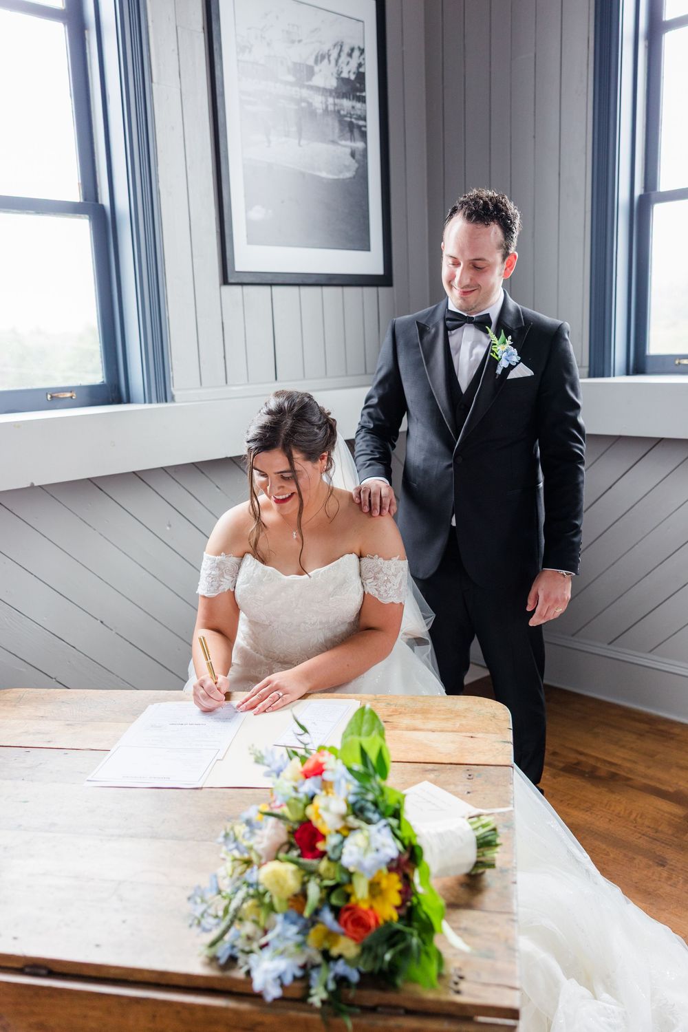 Bride signs wedding documents at wooden table while groom stands beside her, colorful bouquet in foreground.