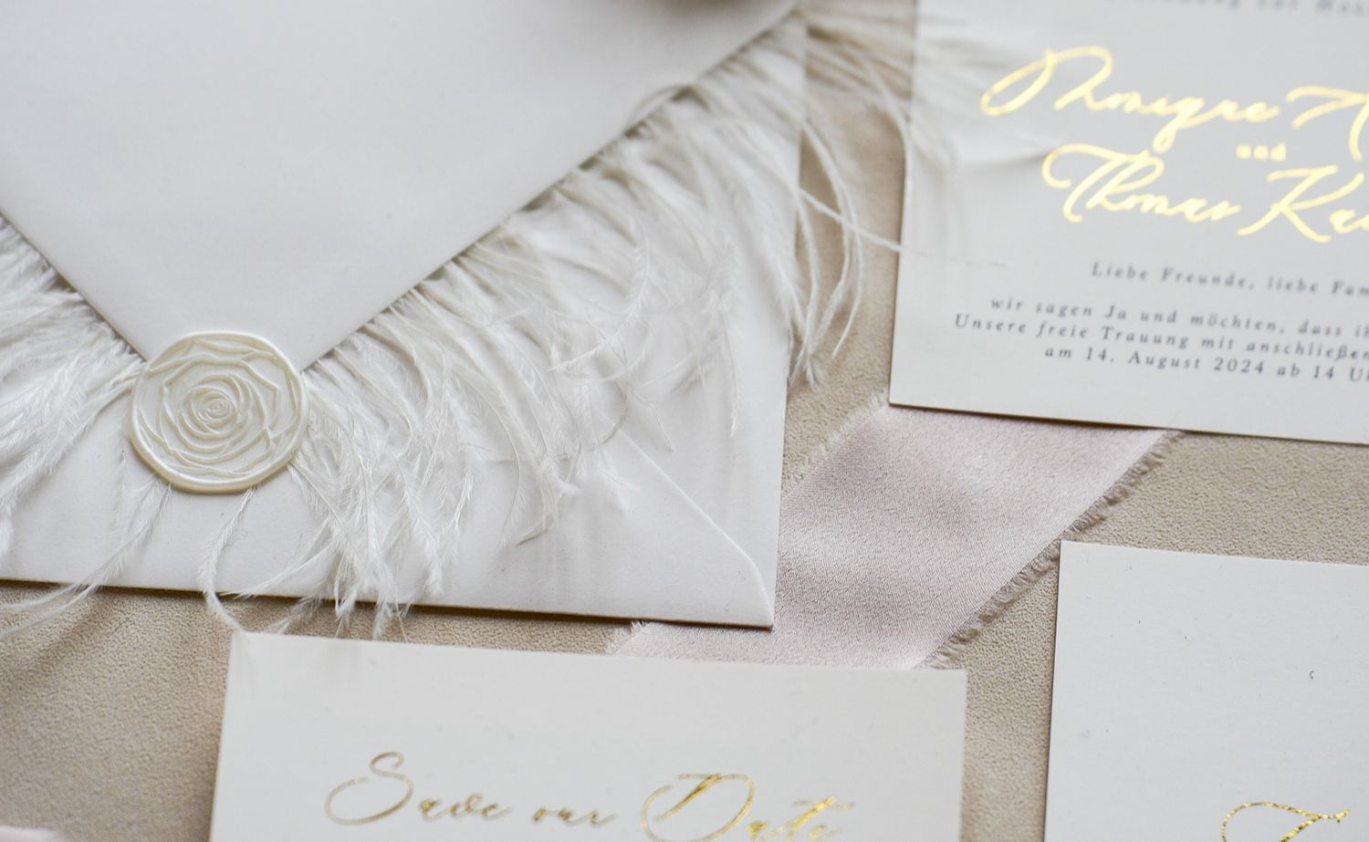 Luxurious wedding invitation suite with gold script, feather details, and wax seal on neutral linen background.