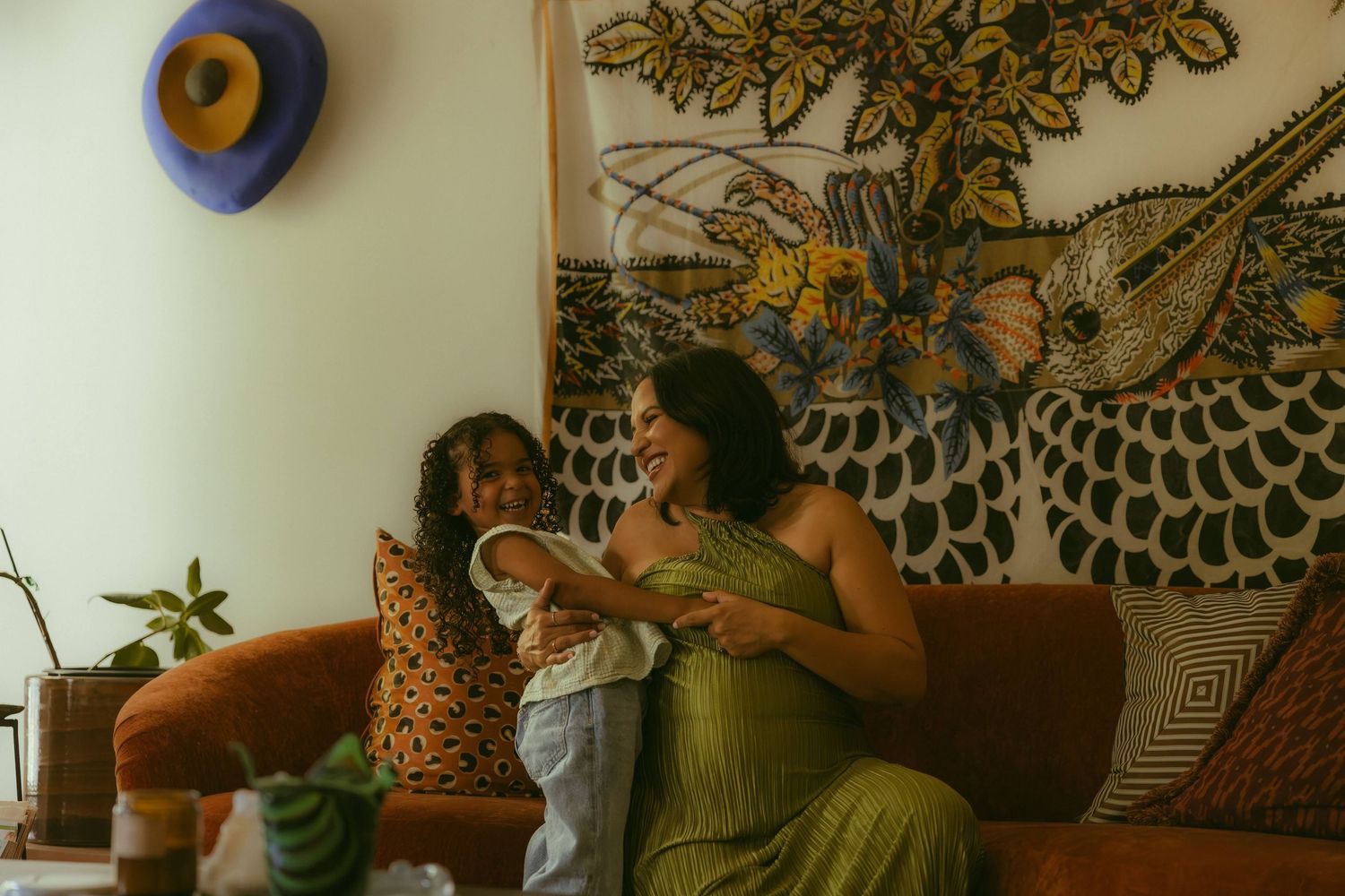 A woman in a green dress sits on a brown sofa smiling with a toddler in a cozy, art-decorated living room during their family photo session.
