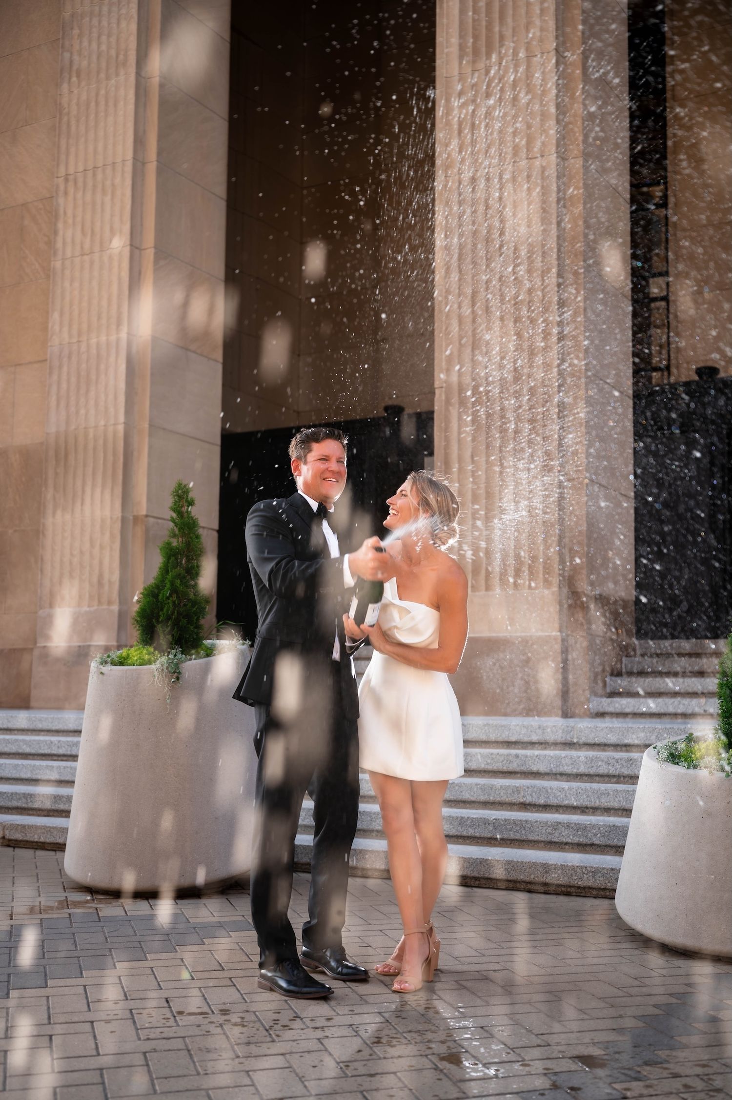 Elegant couple in wedding attire walking hand in hand, woman in white mini dress, man in black tuxedo, urban backdrop.