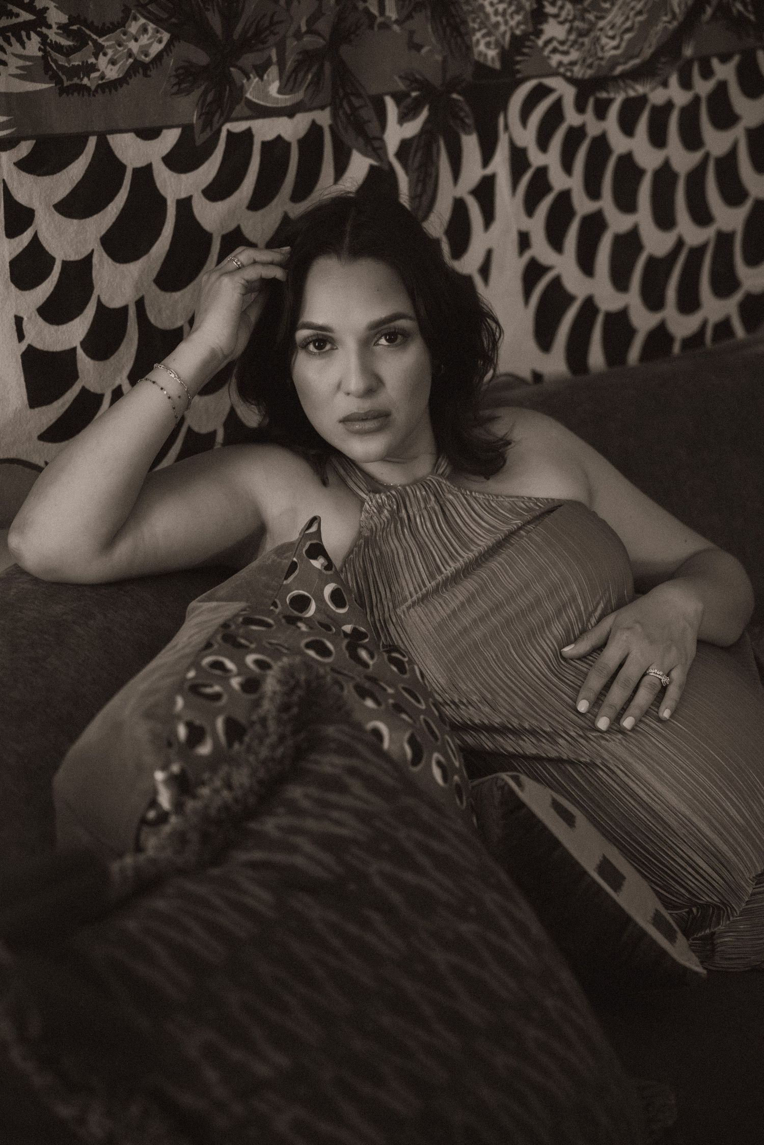 Black and white photo of a woman reclining on a sofa, wearing a pleated dress, with patterned wallpaper behind her.