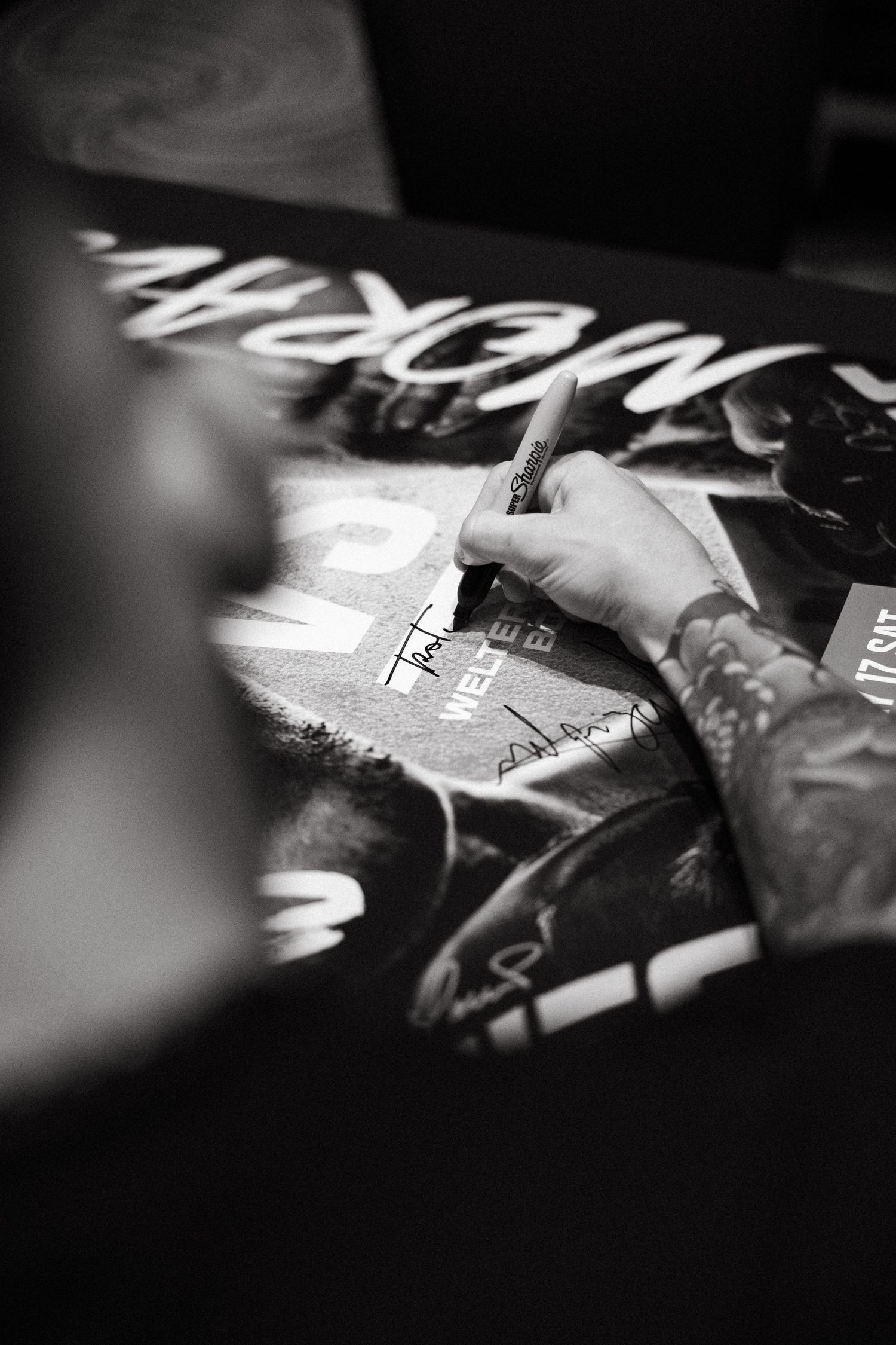 Tattooed hand signing autograph on black and white poster at fan event.