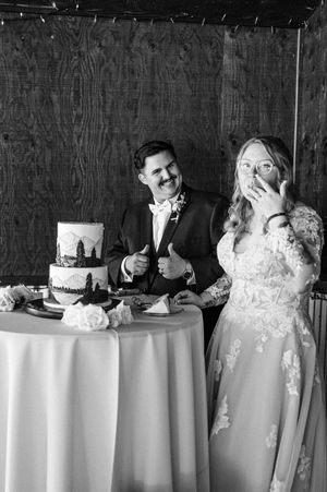 Black and white photo of bride and groom laughing while cutting their decorated wedding cake at reception.