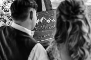 Bride and groom viewing rustic wooden wedding sign reading The Ferences, July 19th 2025 with mountain design.