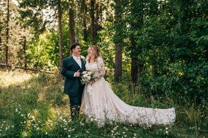 A bride and groom share a romantic moment in a lush forest meadow, surrounded by wildflowers and tall pine trees.