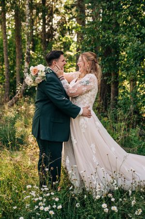 A joyful bride and groom embrace in a wildflower meadow surrounded by lush green trees on their wedding day.
