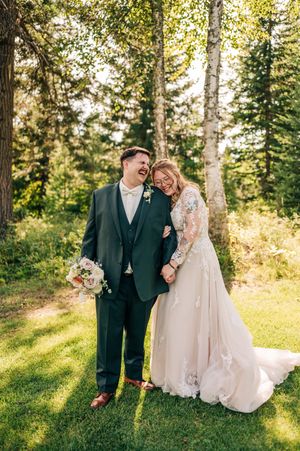 Laughing bride and groom share a joyful moment outdoors, she in lace gown holding bouquet, he in dark green suit.