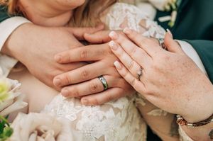 Close-up of bride and groom hands showing wedding rings, bride wearing lace dress and bracelet.