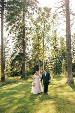 Bride and groom walk hand-in-hand through a sunlit forest clearing on their wedding day.