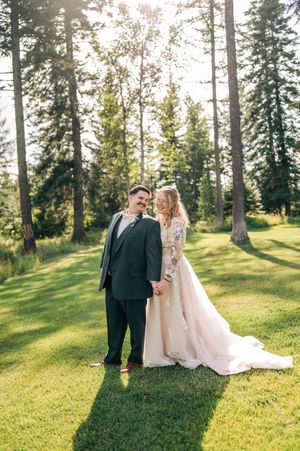 Bride and groom holding hands on lush green lawn surrounded by tall pine trees in golden sunlight.
