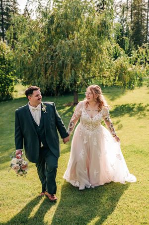 Smiling newlyweds hold hands and walk across green lawn, bride in floral lace gown, groom in dark suit.