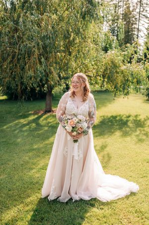 Bride in blush lace long-sleeve wedding gown holding peach floral bouquet standing on green lawn near willow tree.