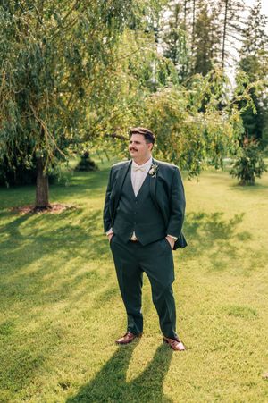 Dapper groom in dark suit with boutonniere stands confidently on green lawn beneath a willow tree.
