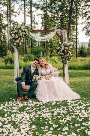 Bride and groom sitting on a vintage sofa under a floral wooden arch surrounded by rose petals on a lush green lawn.