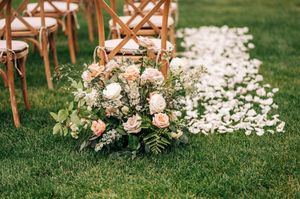 Lush peach and white floral arrangement on green grass beside white rose petal aisle at outdoor wedding ceremony.