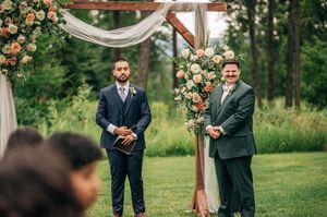 Two men in suits stand at an outdoor wedding ceremony altar adorned with peach and white floral arrangements.