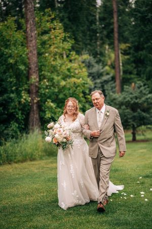 Bride in lace gown walks arm-in-arm with father across green lawn holding peach bouquet at outdoor wedding.