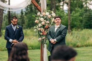 Groom in green suit stands at floral wedding arch outdoors, with officiant in navy suit beside him.