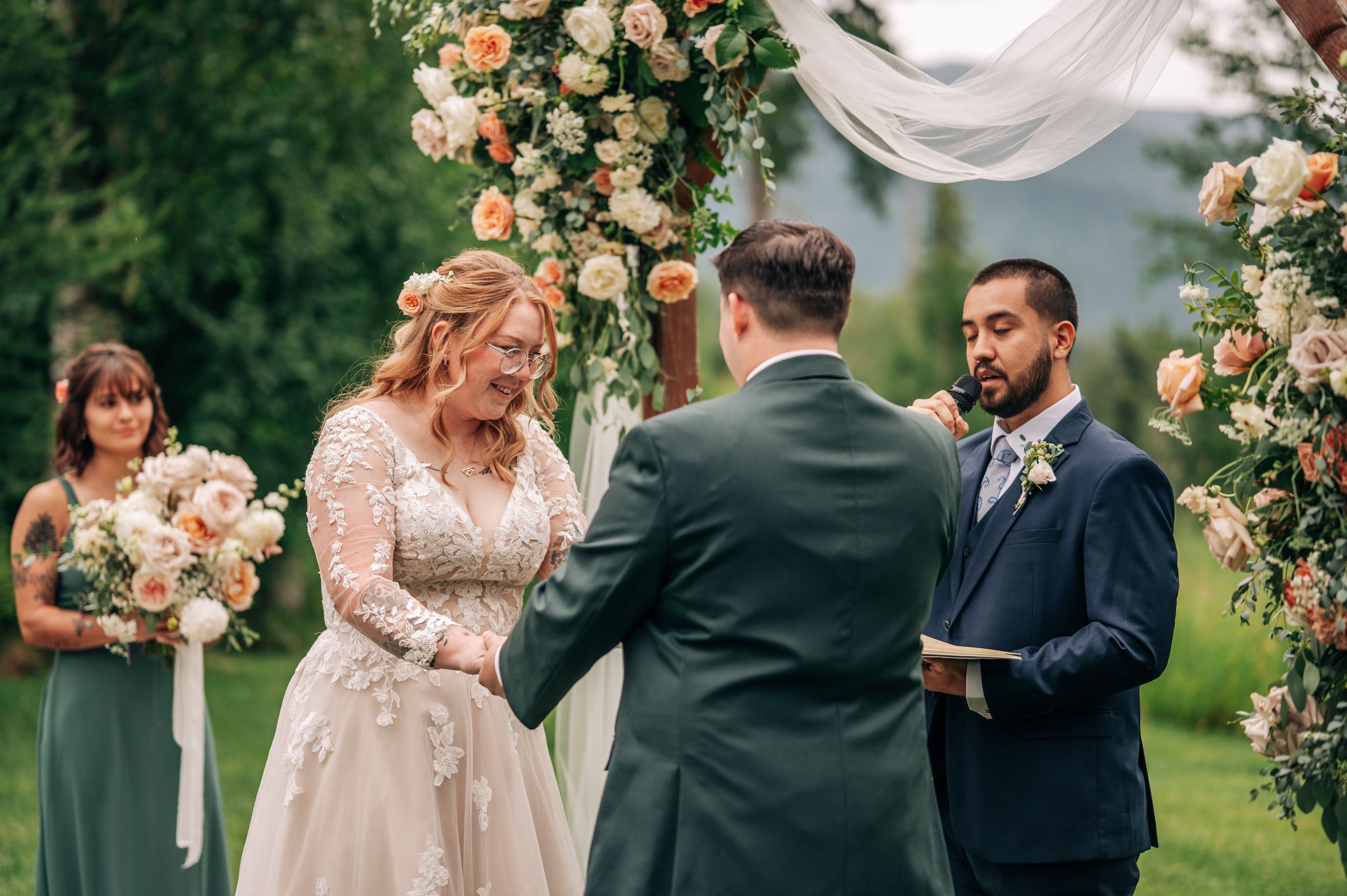 Bride and groom exchange vows outdoors under a floral arch with peach roses, officiant beside them, bridesmaid in sage green.
