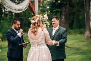 Outdoor wedding ceremony with bride in lace gown, groom in green suit, and officiant reading vows in a garden setting.
