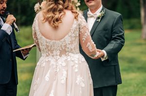 Bride in lace long-sleeve wedding gown holds hands with groom in dark suit during outdoor wedding ceremony.