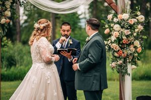 Bride and groom exchanging vows with officiant under a floral wooden arch at an outdoor wedding ceremony.