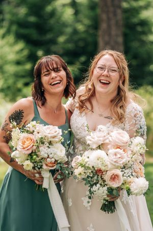 Bride and bridesmaid laughing together outdoors holding beautiful blush and white floral bouquets at a summer wedding.