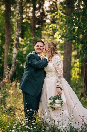 Bride and groom embrace in a sunlit forest, she holds a white bouquet in a lace gown as he smiles in a dark suit.