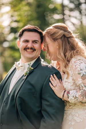 Smiling groom in dark green suit with bride in lace wedding dress sharing a joyful moment outdoors.