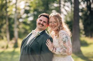 Happy bride and groom posing outdoors on their wedding day, surrounded by lush green trees in natural sunlight.