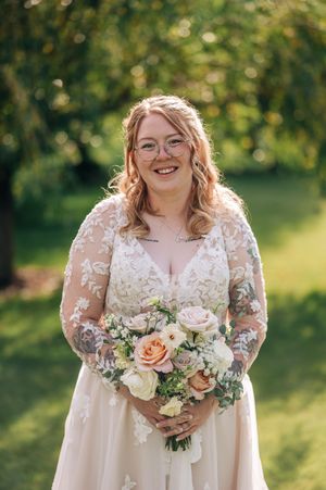Smiling bride in lace long-sleeve wedding gown holding peach and white rose bouquet outdoors.