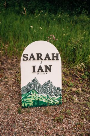 Outdoor wedding sign for Sarah and Ian dated 07.19.25 featuring painted mountain scenery on white arch board.